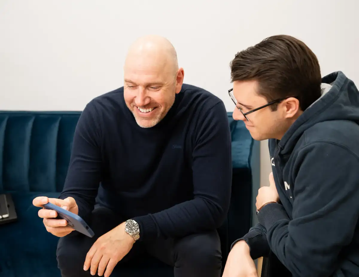 Zwei Männer auf einem blauen Sofa schauen lachend auf ein Smartphone